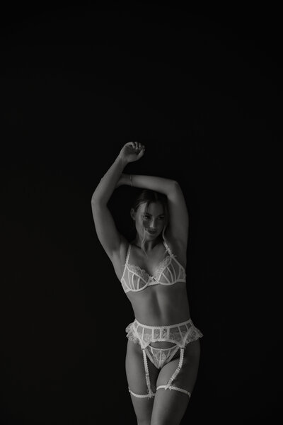 Black and white photo of woman in lingerie with her arms above her head standing in front of a black wall
