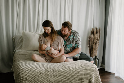 Mom wearing tan dress sitting next to dad while holding baby and sitting on a cream bed in studio in Annapolis Maryland photographed by Bethany Simms Photography