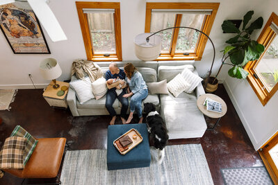 birds-eye view of dad holding newborn on couch next to mom with dog looking on