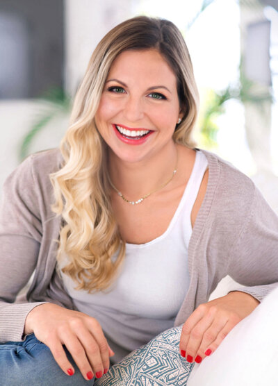 A portrait of East Coast wedding photographer Anna Schmidt. She has long, wavy blonde hair and is sitting indoors, smiling. She wears a beige cardigan over a white top, with her hands resting on a cushion with a geometric pattern