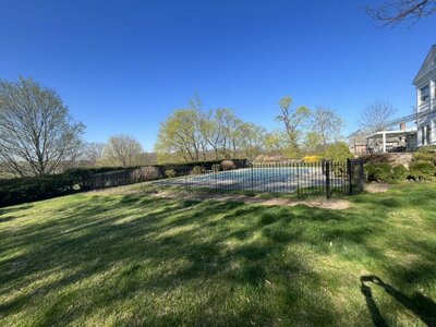 A backyard with a grassy lawn and a fenced-in pool area before landscaping, with a clear blue sky and trees in the background.