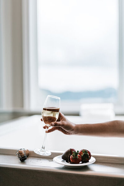 Hand holding a glass of champagne in a spa bath with chocolate covered strawberries on a plate