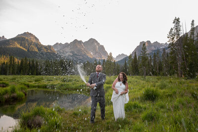 Bride and groom pop champagne in an alpine meadow, celebrating with mountain peaks in the background.