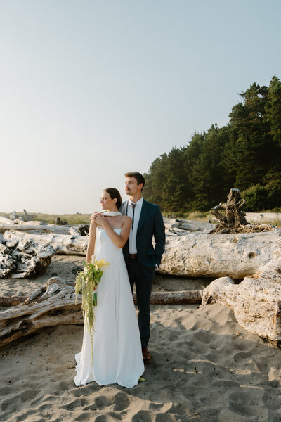 A couple poses for portraits at sunset in front of driftwood during their oregon coast wedding. 