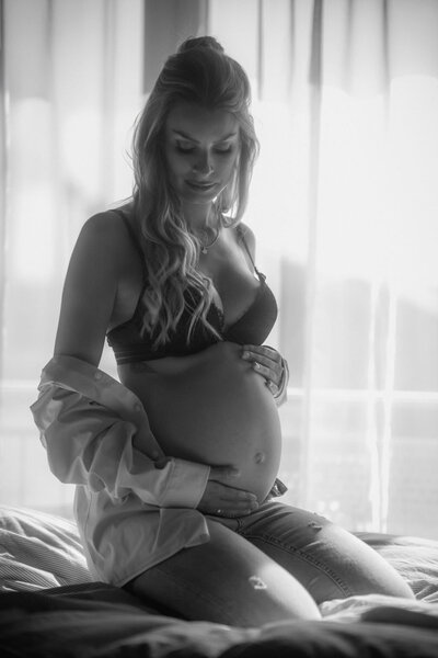 Eine schwangere Frau auf einem Bett kniend, die ihren Babybauch hält und zur Seite schaut.