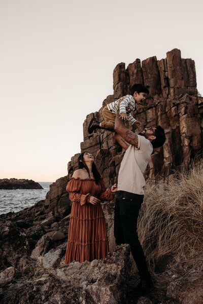 A man is lifting a child into the air while a woman watches. They are standing on rocky terrain near the sea, with large rock formations in the background. Perfect for Wollongong family photography, this picturesque scene captures the essence of outdoor family sessions in Illawarra.