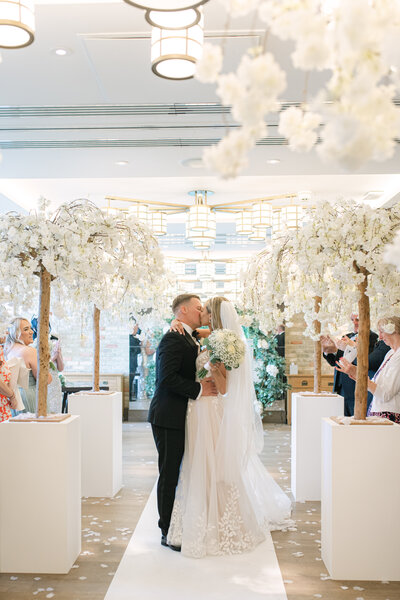 Bride and Groom are standing in front of Syon Park , looking back over their shoulder towards the camera. The bride is wearing a white lace dress with a long veil and the groom is in a black tux