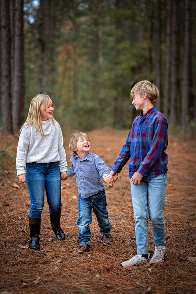 A sister is holding hands with her two brothers while they walk through a forest while they take family pictures.