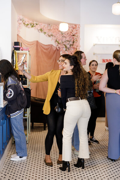 A woman in a yellow blazer and another in a black off-the-shoulder top pose for a selfie at a stylish café during a networking event in Seattle. Other patrons are engaged in conversation near the counter, with pink floral decor and modern light fixtures in the background.