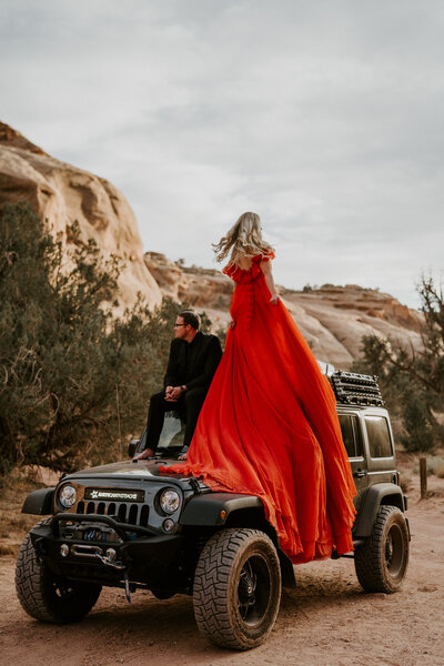 Woman in dramatic red dress stands on Jeep as man sits nearby, adventure elopement photo near Breckenridge, Colorado.