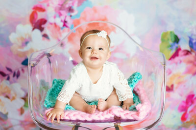 Milestone photographer,  a young baby sits on a clear chair with a colorful background