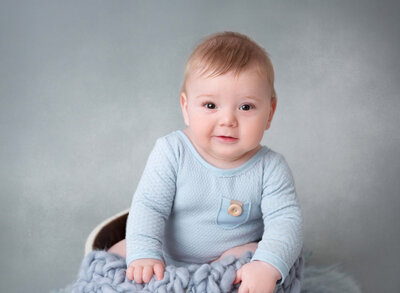 First birthday portrait by top Brooklyn and NYC Baby Photographer Rochel Konik. Baby is witting in a white knit long-sleeved onesie with wooden toys stacked in front of him. Baby is looking at the camera and smiling.