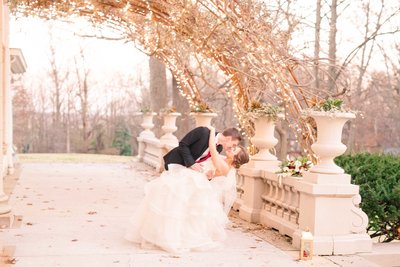 A bride and groom dip into a romantic kiss under the christmas lights of the Liriodendron Mansion for a Christmas wedding