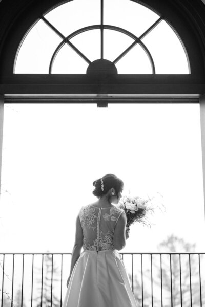 La mariée respire délicatement le parfum de son bouquet devant une fenêtre baignée de lumière, moment paisible et raffiné capturé lors des préparatifs d’un mariage élégant