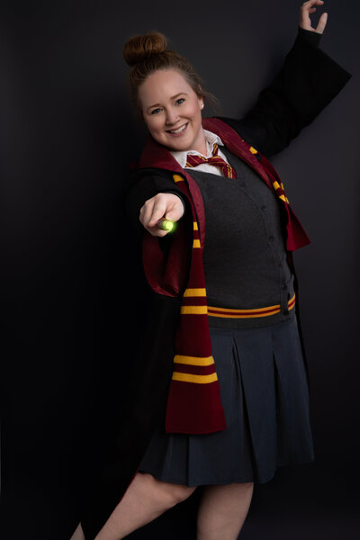 Woman waving wand dressed in Harry Potter costume with scarf, cape, and necktie. Wand has a green glow at tip.