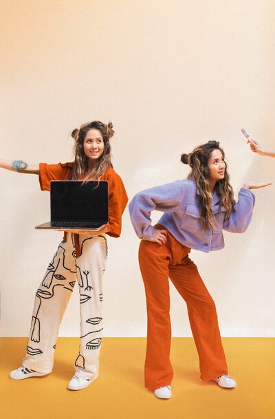 Woman with half-up pigtail buns holds a laptop wearing abstract face printed pants standing next to a photoshopped duplicate version of herself wearing a purple top and orange pants while posing for a picture