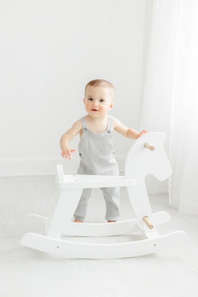 One year old baby boy wears overalls and stands next to white rocking horse during portrait session