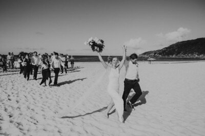 Wedding photograph of a bride jumping for joy as she leaves her ceremony on the beach with her new husband. Natural and relaxed. Central Coast beach wedding.