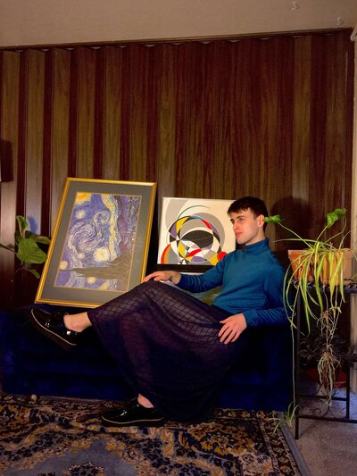 Carter Jones  sits on a couch, surrounded by artwork and plants, against a wood-paneled wall. Representing his vendor profile for the Crush Collective wedding show.