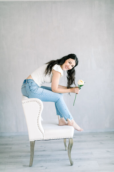 Brunette headshot brand photos on White linen chair, smiling and holding a flower.