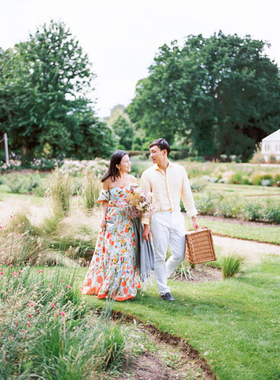 engaged couple walking at the chiswick gardens while bride to be holding flower bouquet and fiance holding picnic basket and blanket