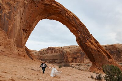 A bride and groom run towards the camera after officially getting married at the Corona Arch in Moab