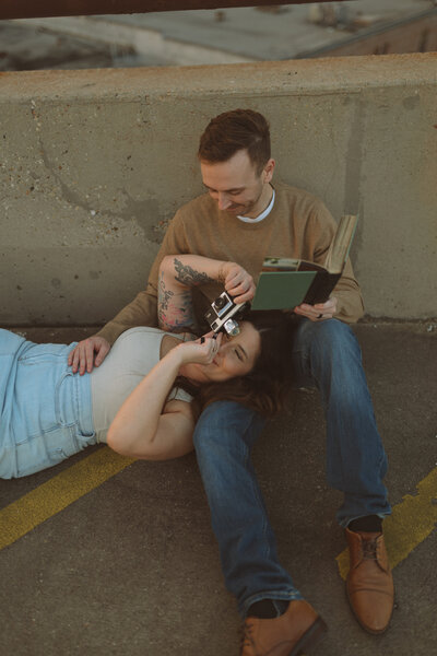 a man reading a book while his wife rests her head on him taking photos through an old vintage film camera.
