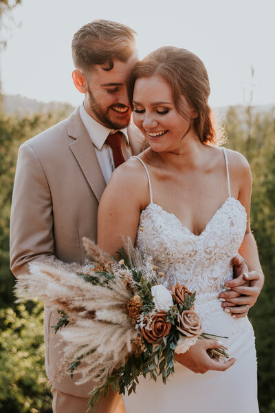 Bride and groom embrace while standing in front of forest sunset