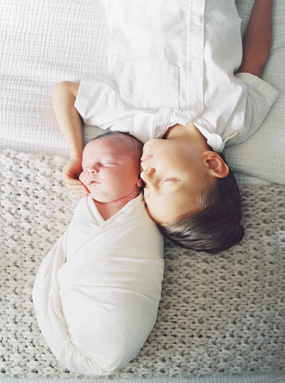 Baby swaddled in white blanket asleep on bed next to his big brother that is snuggling him, both eyes closed. 