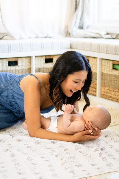 A mother smiles at her baby during tummy time in a bright home nursery, a heartwarming moment by Ellobelle Photography.