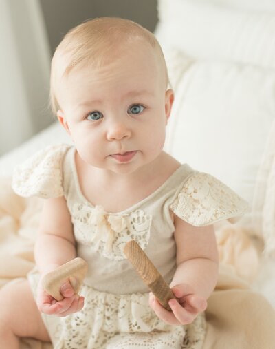 Baby in a delicate lace outfit sits on a cozy bed, holding a wooden toy and gazing softly. A timeless baby session by Moments in Light Photography.