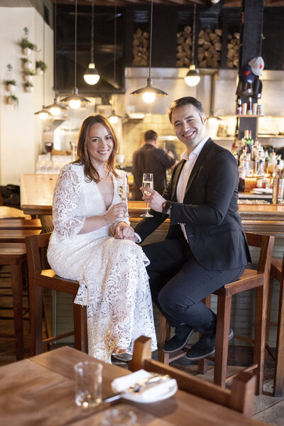 A couple sits at a bar, dressed in formal attire. The woman wears a white lace dress, and the man wears a black suit. They are smiling and toasting with champagne glasses. The background shows bar shelves and warm lighting.