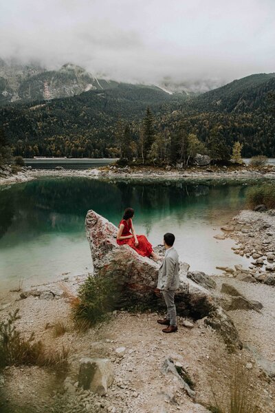 An eloping couple wearing a red dress and grey suit looks out  over Eibsee near Garmisch