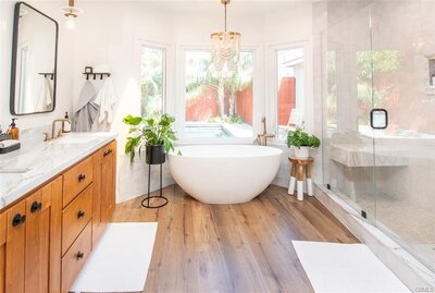 Bright master bathroom with soaking tub, bay window, chandelier, hardwood floors, white walls, and potted ferns for a serene, elevated feel.