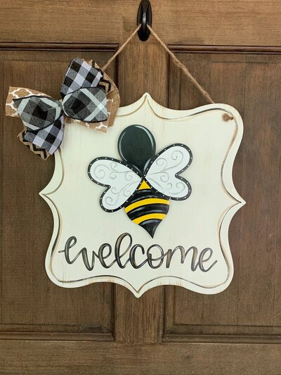 white rectangle wooden door hanger with bumble bee and black hand lettered welcome
