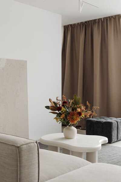 A serene, minimalist living room featuring a white coffee table with a glass vase of fresh flowers. A beige sofa sits in front, while soft brown curtains create a warm, inviting ambiance. Styled for Bloom Botanicals’ Instagram strategy, this image embodies a modern, organic aesthetic.