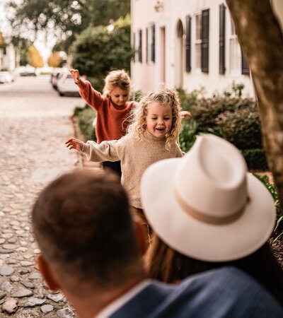 Playful children running around on a cobblestone street in Charleston while parents watch with joy in a candid lifestyle family session.