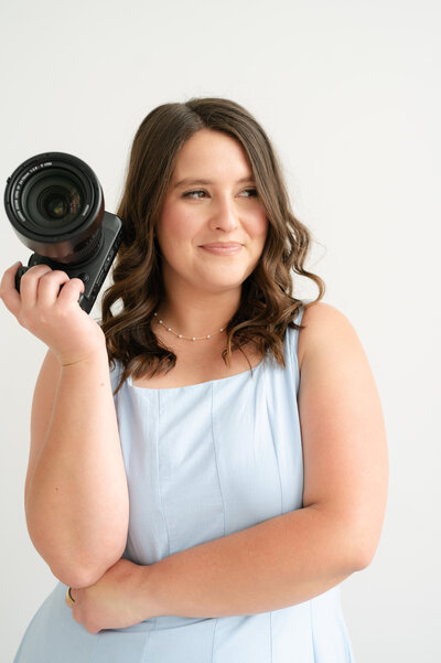 Wedding Photographers in Rhode Island | Kelsey Sheehan Photography Timeless Rhode Island Weddings | Kelsey holding a camera, smiling slightly. She wears a light blue sleeveless dress, against a plain white background.