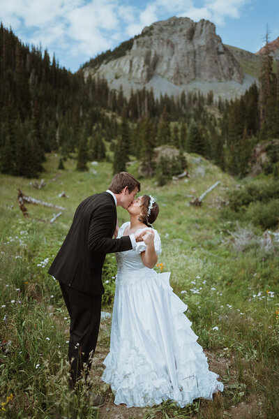 Couple standing out opposite doors of their jeep cheering and smiling at each other. the bride is holding her bouquet, and the groom is wearing a black suit. the jeep is silver. they are in the colorado mountains after their hiking elopement.