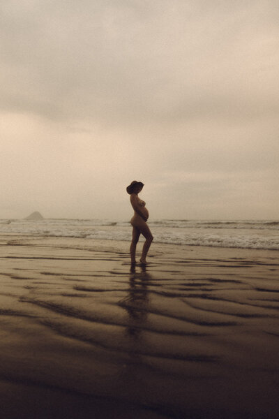 A portrait of a pregnant naked woman holding her belly on the sand of Ohope beach. Captured by Eilish Burt Photography