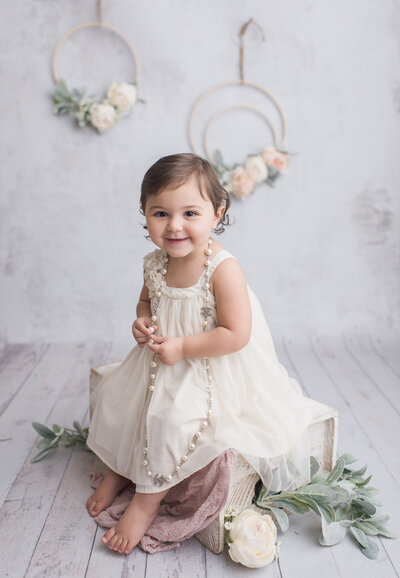 a 3-12 month old baby sitting on a stool with a white dress on