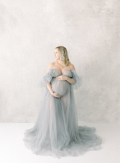 A expecting mom, standing inside the studio of a las vegas maternity newborn photographers space in front of the window looking out while she is wearing a tull gown that the photographer provided.she chose the gown from miss rose photography client wardrobe collection of gowns.