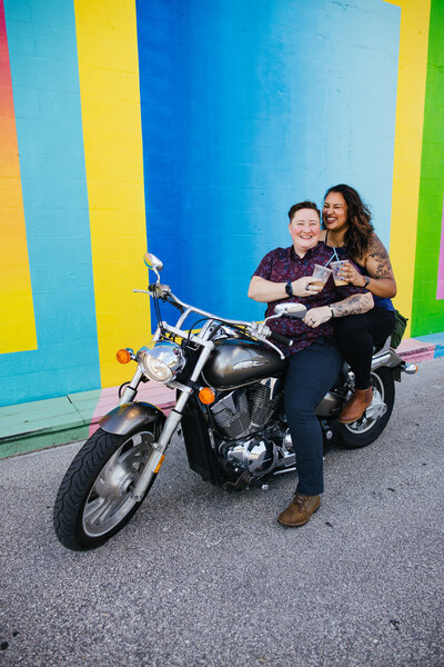Two women on a motorcycle in front of a colorful mural for their Engagement Photos
