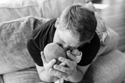 A black and white image of a father sitting on a couch, cradling a newborn in his hands and kissing the baby's head tenderly, capturing a quiet moment of affection.