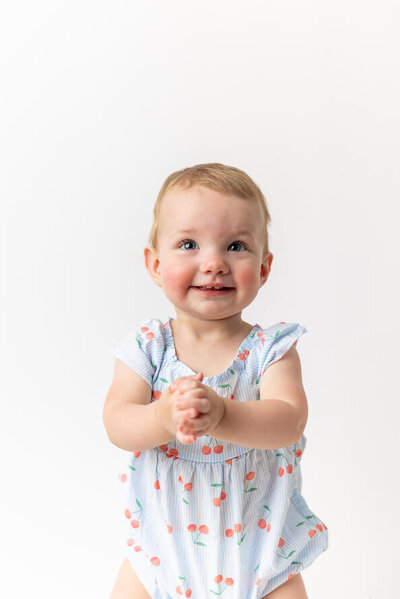 Baby girl smiles at camera and clasps hands in a white photo studio.
