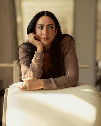 A woman with long dark hair leans on a white chair, resting her chin on her hand and gazing thoughtfully to the side. She wears a brown sweater and sits in a softly lit, neutral-toned room.