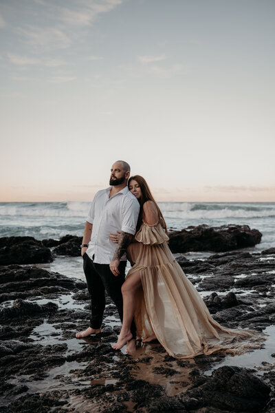 A woman in a sheer white dress stands barefoot on a rocky beach at sunset, looking out towards the water, capturing the serene beauty perfect for Shellharbour maternity portraits.