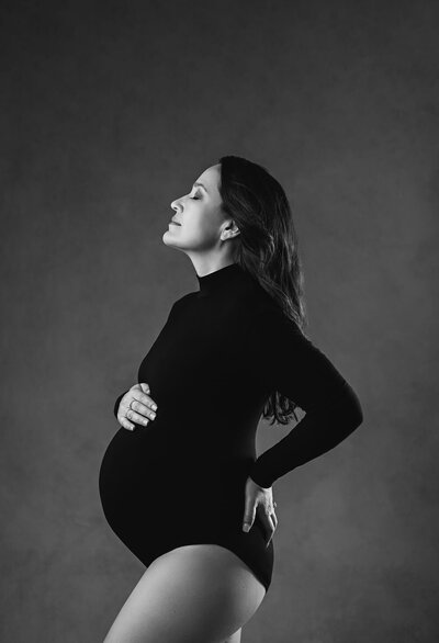 Artistic portrait in black and white where  pregnant woman is wearing a bodysuit an looking up showing passion while holding her bump