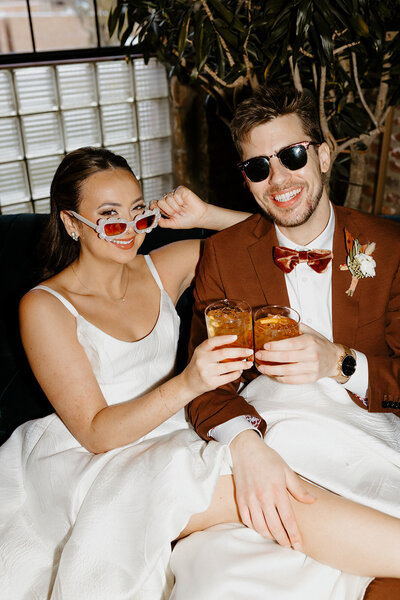 Bride and groom playfully cheers and smile at camera wearing sunglasses at their reception.