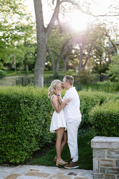 A couple stands closely next to a tree. The man gently touches the woman's chin. They exchange warm, affectionate gazes in a serene, outdoor setting.
